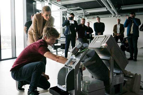 En studerende fra Syddansk Universitet i Sønderborg viser et produkt, som Statsminister Mette Frederiksen kigger på. I baggrunden står blandt andre en gruppe fotografer og tager billeder. 