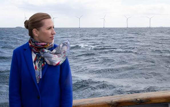 Statsminister Mette Frederiksen står på en båd - med havet og vindmøller i baggrunden.
