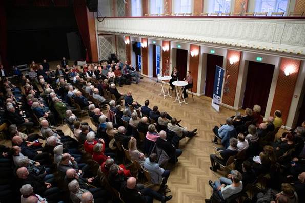En sal fyldt med publikum, der ser i retning af statsminister Mette Frederiksen, som sidder ved et højt rundbord. 